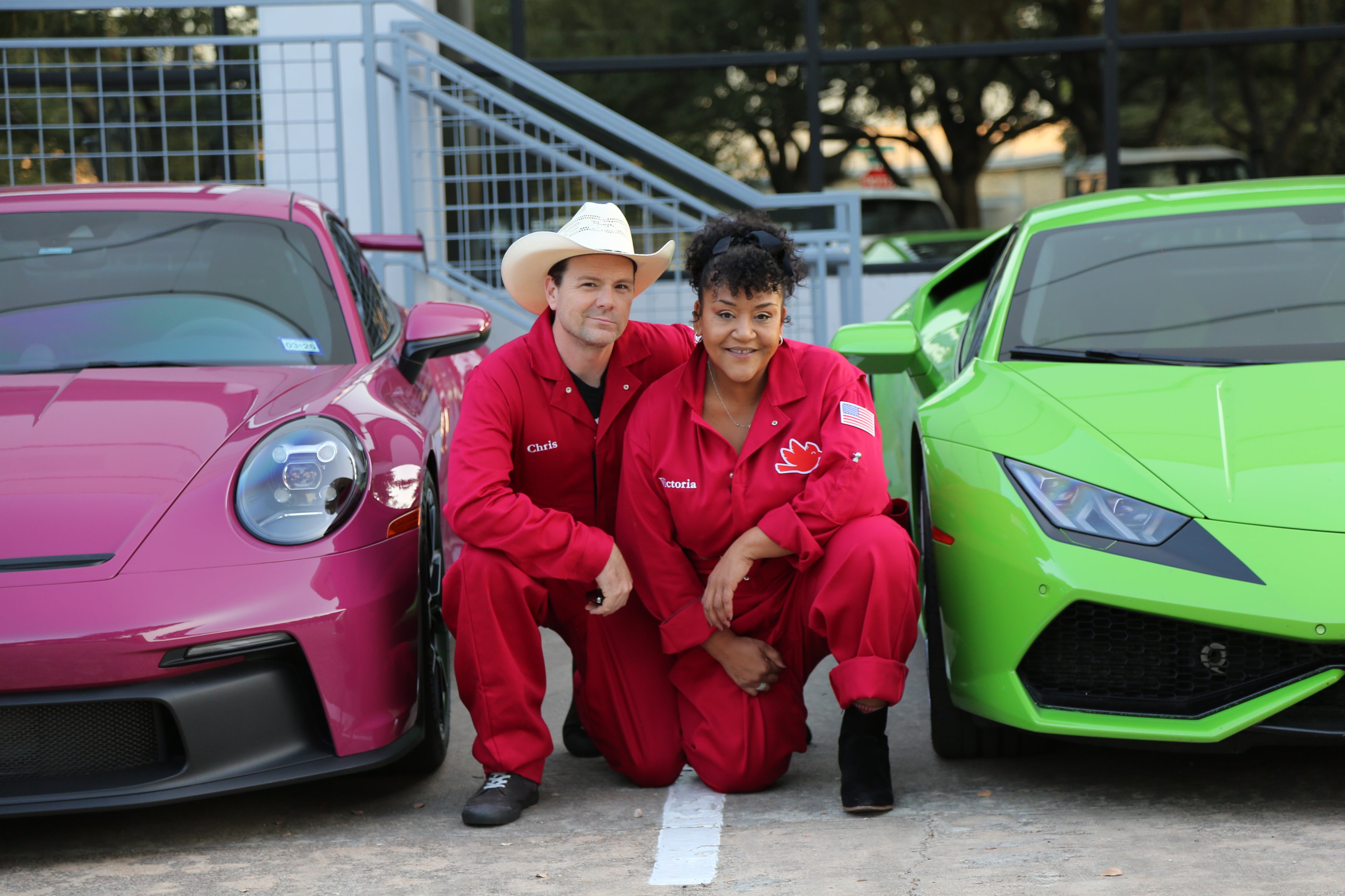 Chris and Victoria Clozure outfits posing between a pink car and a green sports car.